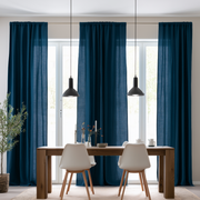 A pair of long navy blue curtain panels hanging in front of tall windows in a bright dining room with a wooden table, white chairs, and black pendant lights.