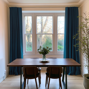 A dining room scene with a wooden table and four chairs in front of a large multi-pane window. Deep teal curtains frame the window, and a vase with flowers sits on the table.