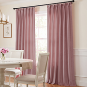 A pair of dusty rose velvet curtain panels hanging on a black rod in a bright dining room with a white table and chairs.