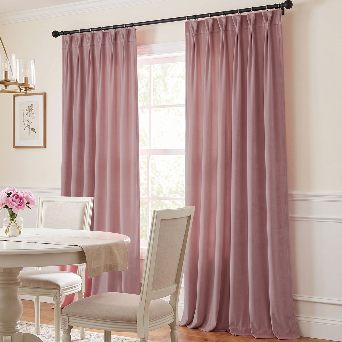 A pair of dusty rose velvet curtain panels hanging on a black rod in a bright dining room with a white table and chairs.