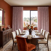 A styled dining room with a dark wood dining table set for six, surrounded by cream upholstered chairs and a prominent rust-orange velvet chair in the foreground. The room features terracotta walls, matching pink pleated curtains, and a large window overlooking a landscape.