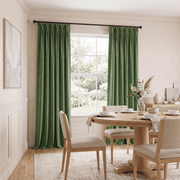 A pair of sage green curtain panels hanging on a black rod over a tall window in a bright, neutral-toned dining room with a wooden table and chairs.