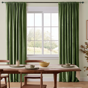 A pair of dark green curtain panels hanging on a black rod over a tall window in a dining room setting with a wooden table and chairs.