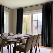 A dining room with tall black curtains hanging on both sides of large windows, with a wooden dining table and upholstered chairs in front.