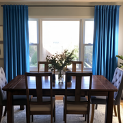A pair of bright blue curtain panels hanging on a black rod in a dining room setting with a wooden dining table and chairs.