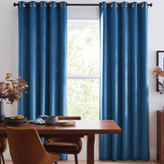 A pair of deep blue grommet-top window curtains hanging on a black curtain rod in a bright dining room setting with a wooden table and chairs.