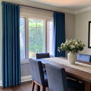A pair of deep blue velvet curtain panels hanging on a rod over a multi-pane window in a dining room setting with a wooden table, blue chairs, and a white vase of flowers.