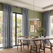 A dining room interior with tall gray curtains hanging around large windows, a round white dining table, wooden chairs, framed wall art, and a hanging pendant light.