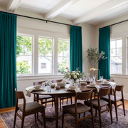 A styled dining room with a round wooden dining table set for six, surrounded by mid-century modern wooden chairs with upholstered seats. The room features high ceilings with white beams, large multi-pane windows, and floor-length teal curtains.
